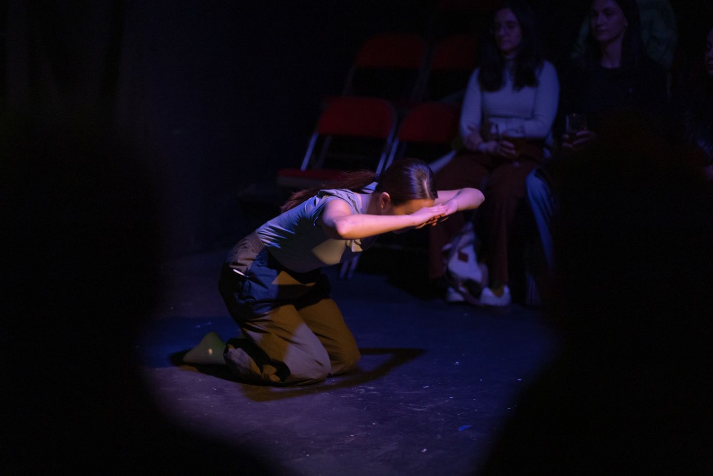 A performer kneels and reaches forward, close to the audience, in warm light.