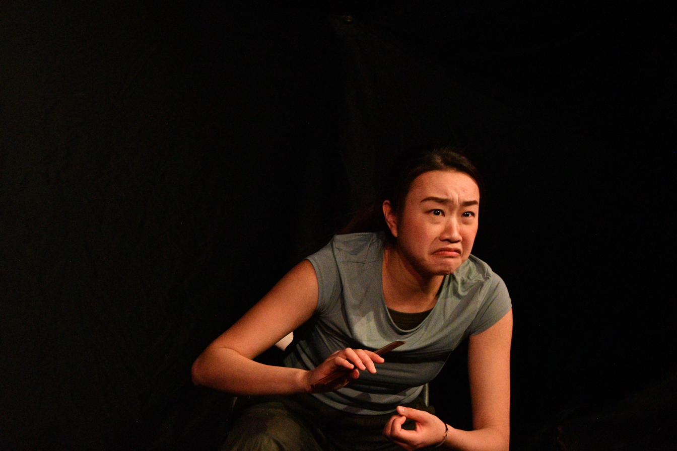 A performer holds a comb, mid-emotion, against darkness in Between Strands.