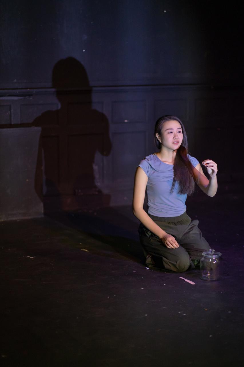 A performer kneels beside a glass jar, her shadow cast large on the wall behind.