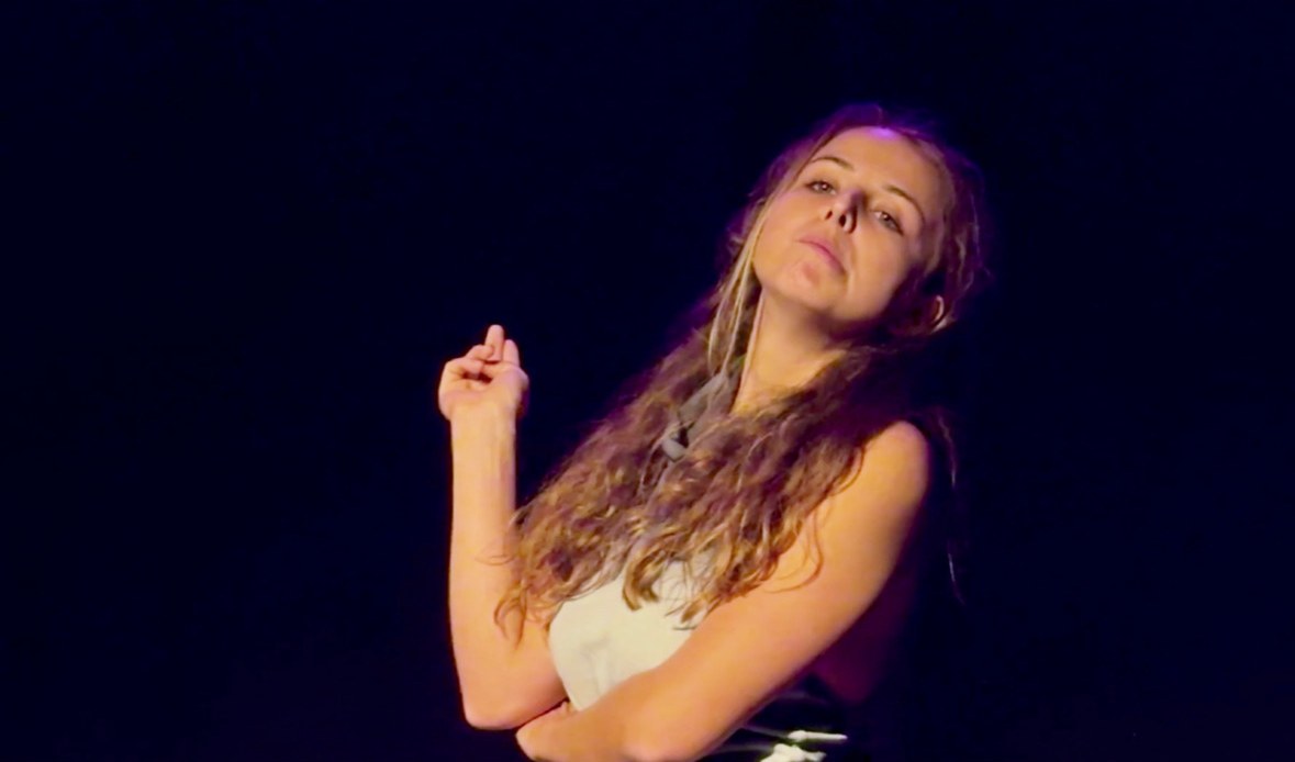 Lalia tilts her head back with a knowing look, one hand lifted.