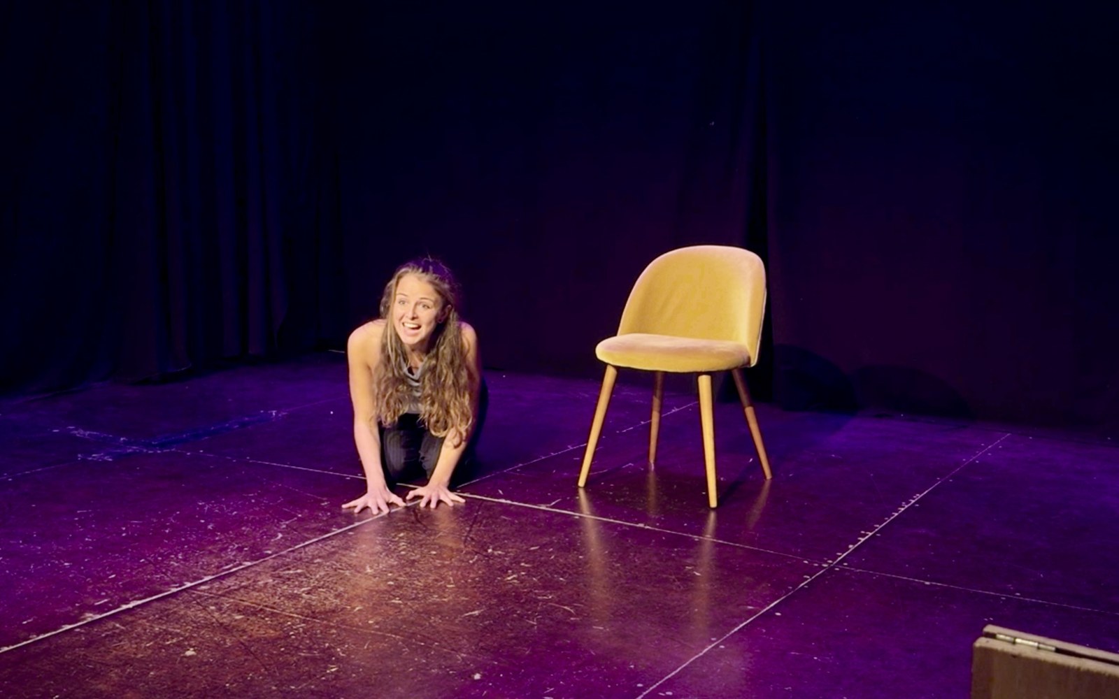 Lalia kneels on the stage floor, leaning forward with a bright, animated expression beside an empty yellow chair.