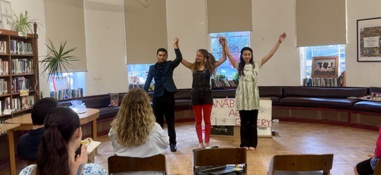 Three performers bow with arms raised after a workshop in a library.
