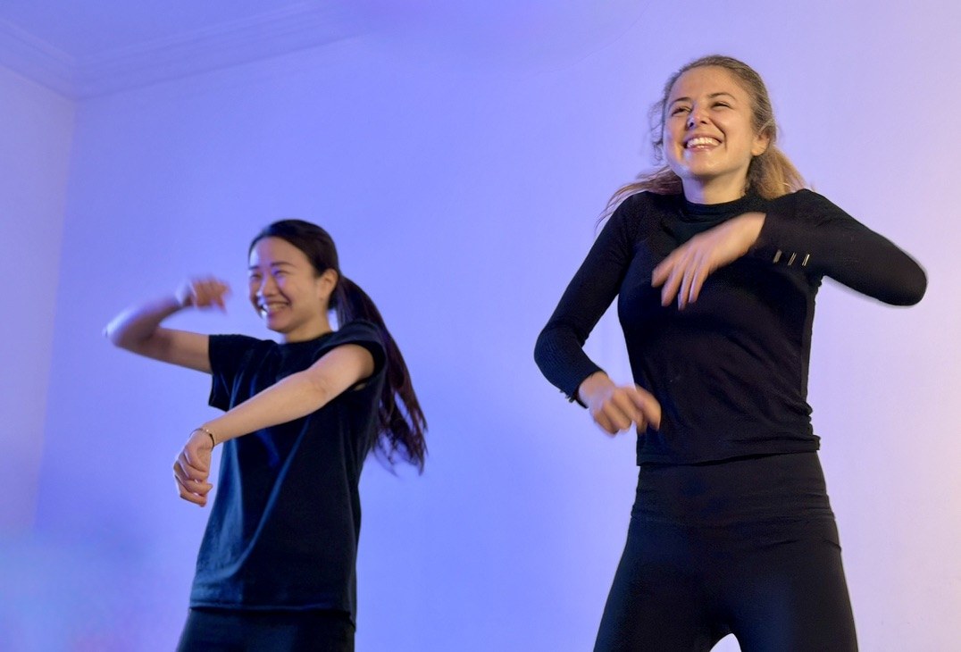 Two performers laughing mid-movement against a violet wall.