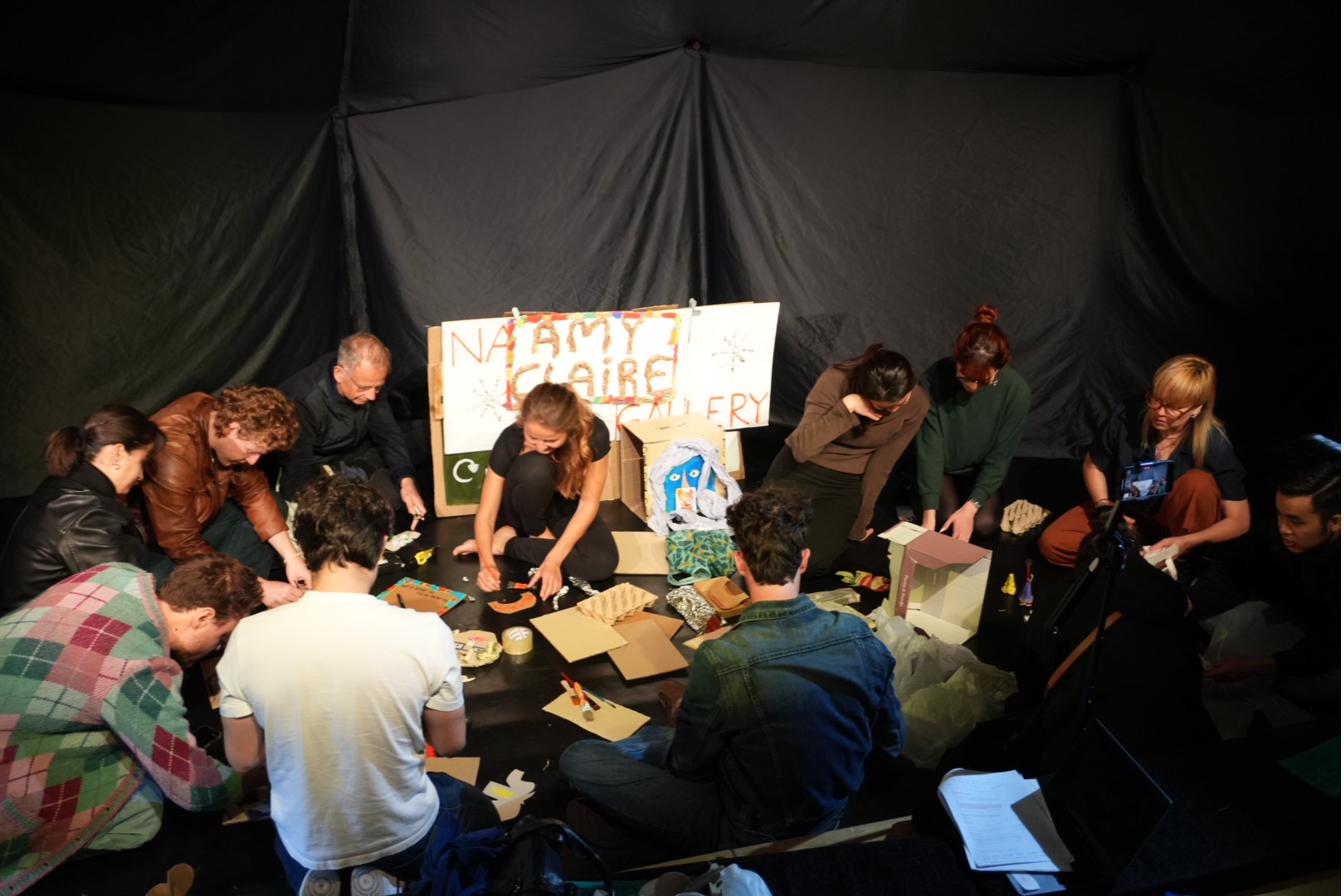 A circle of participants make art from cardboard and waste around a hand-painted gallery sign.